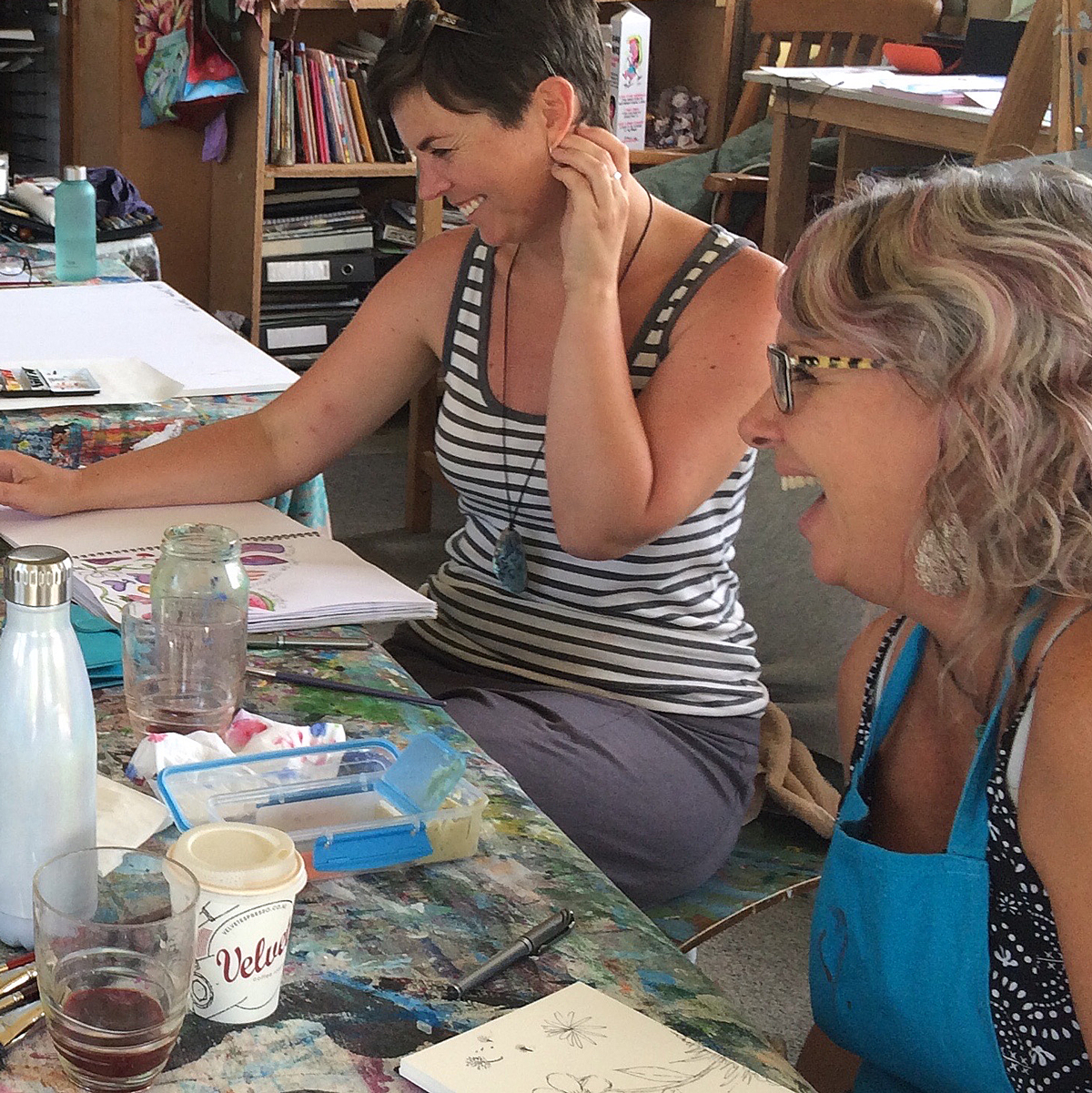 Two women laughing at a paint-stained work table in a studio, sketchbooks open and brushes in hand