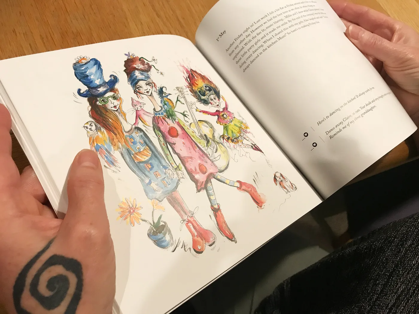 Claire's open journal held in two hands on a wooden table, her watercolour illustration of three dancing figures facing a handwritten entry